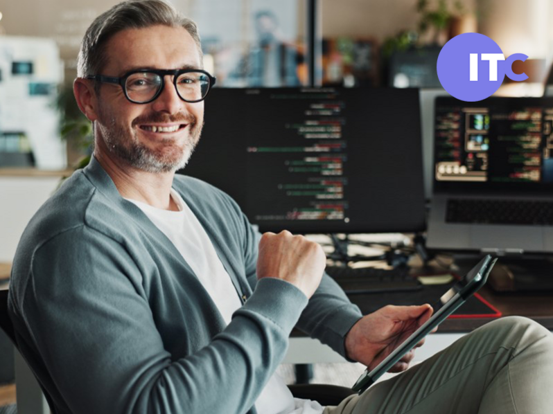 Cyber-Security-Management (m/w/d) Softwareentwickler sitzt am Schreibtisch und lächelt in die Kamera. Im Hintergrund Bildschirme mit offenen Anwendungen.