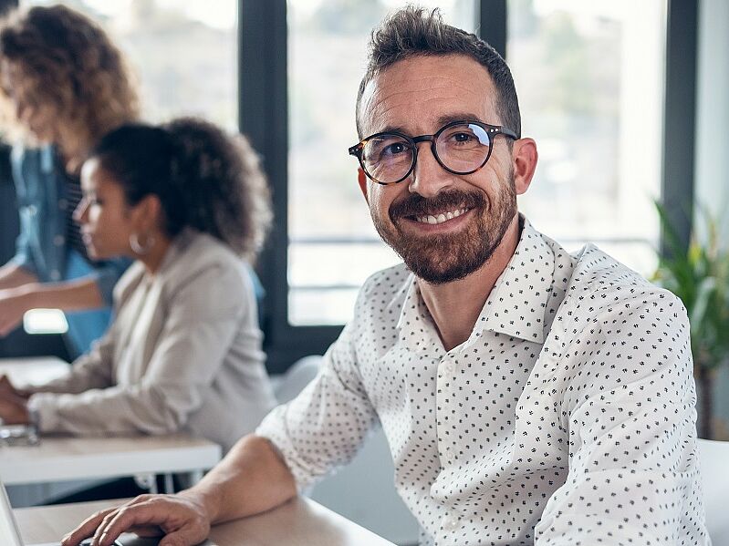 Sachbearbeiter:in Biltrolling (m/w/d) Freundlicher Sachbearbeiter sitzt am Arbeitsplatz und blickt lächelnd in die Kamera