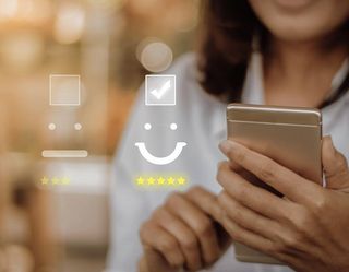 Frau hält ein Handy in der Hand. Links im Bild sind drei Smileys mit Ankreuzkästchen zu sehen: ein trauriges, ein neutrales und ein glückliches. Der Haken ist beim glücklichen Smiley gesetzt.