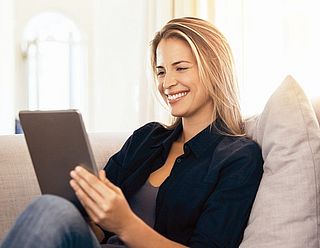 Frau bedient ein Tablet und sitzt dabei auf einer Couch