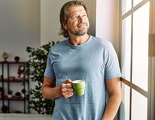 Mann hält Kaffeetasse in der Hand und schaut aus dem Fenster