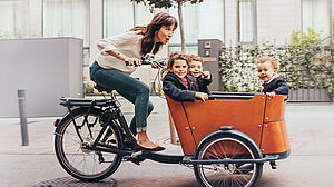 Drei Kinder sitzen vorne in einem Aufsitzer, während die Frau mit dem Fahrrad fährt.