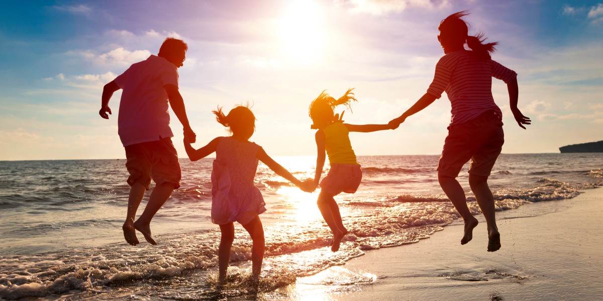 Familie am Strand springt Hand in Hand in die Luft. Im Hintergrund sind das Meer und der Sonnenuntergang zu sehen