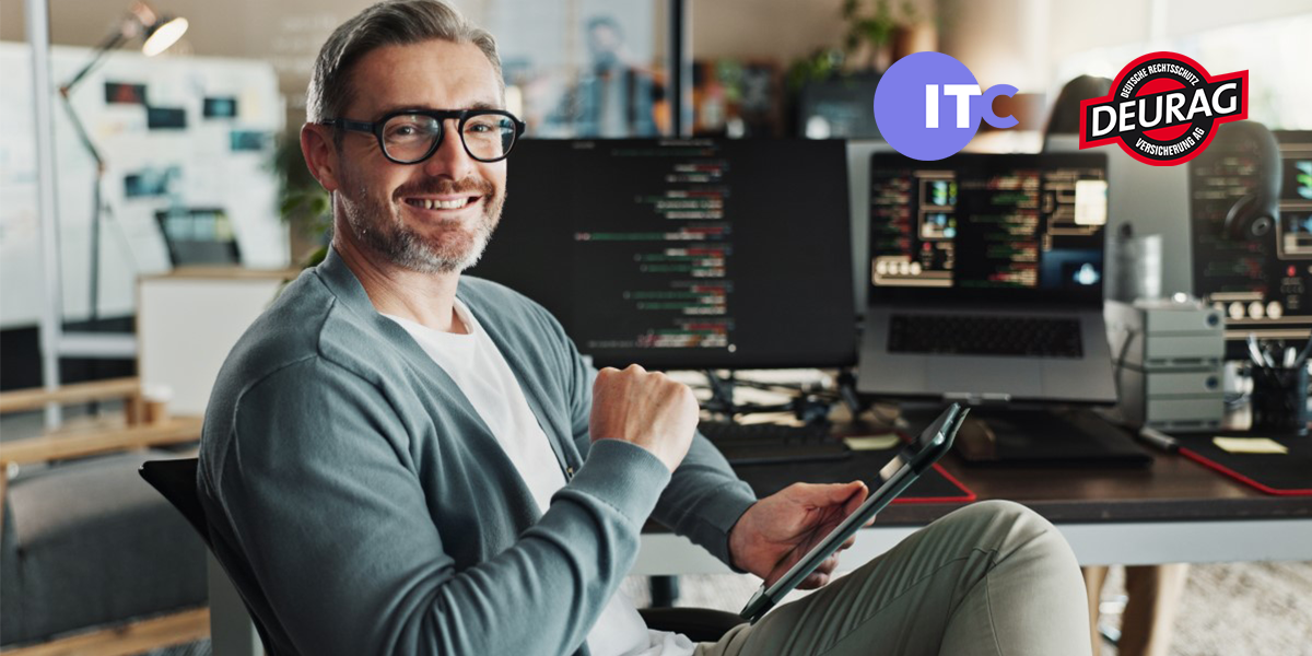 Softwareentwickler sitzt am Schreibtisch und lächelt in die Kamera. Im Hintergrund Bildschirme mit offenen Anwendungen.